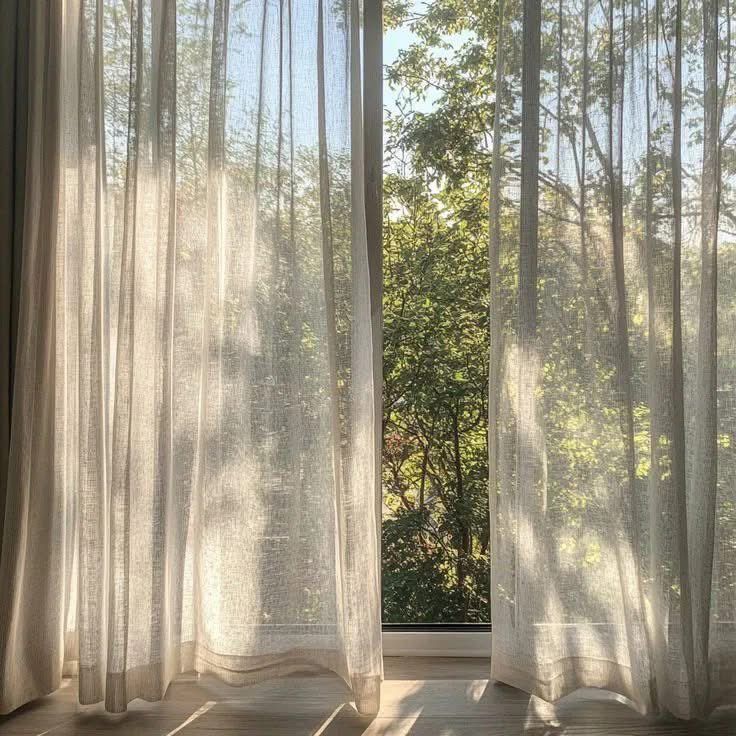Sheer linen curtains drawn back from a window full of green trees and morning sun